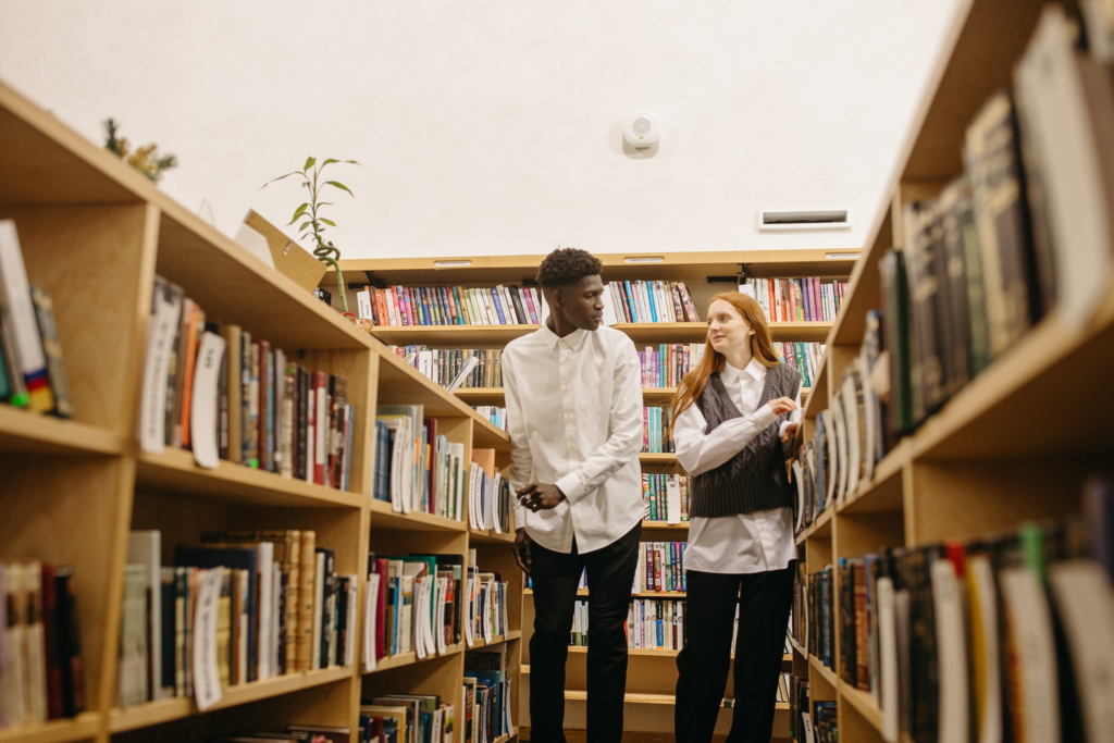 Twee studenten staan tussen twee rijen boekenkasten. Welkom.